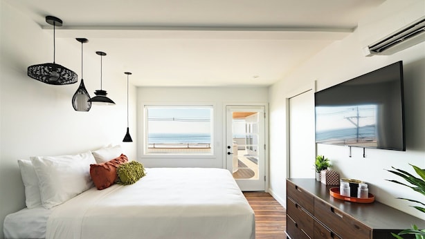 A modern bedroom with a large bed, a TV mounted on the wall, and a view of the ocean through a door. - The Nevada (York Beach)