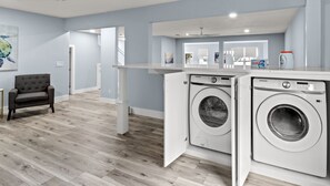 A laundry room with two Samsung washing machines, a grey upholstered chair, and a white countertop with various items. - #1 Destin home, Saltwater spa & pool, Beach@2min walk, Golf cart (Destin)