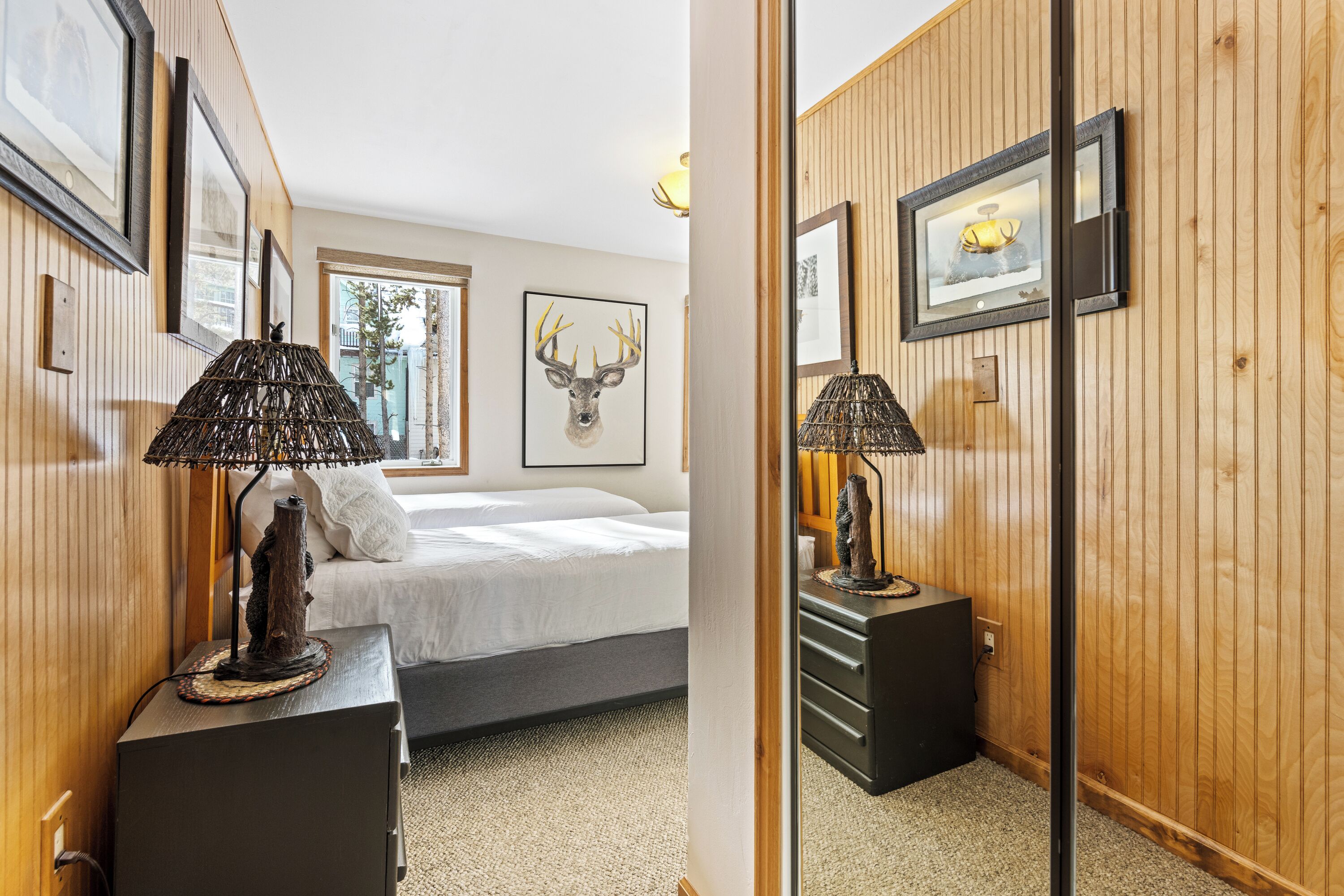 A hotel room with a bed, a lamp, a framed deer head artwork, and wooden paneling.