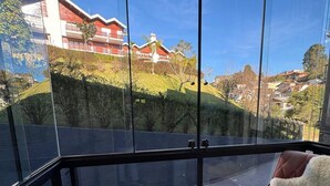A modern room with a large glass window, a plush sofa, and a view of a hillside with buildings. - Loft cama Kingsize 3min centro turístico (Campos do Jordão)