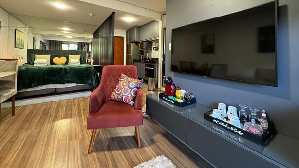 A modern hotel room with a bed, a red armchair, a TV, and a minibar. - Loft cama Kingsize 3min centro turístico (Campos do Jordão)