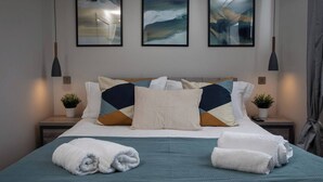 A neatly made bed with a teal bedspread, multiple pillows, and two bedside tables with small potted plants.