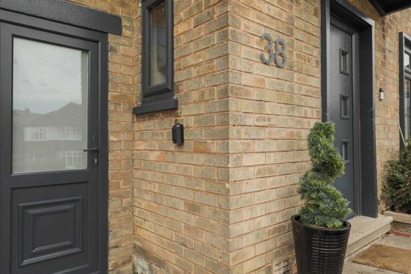 A brick building with a number '38' on the wall, a black door, and a potted plant.