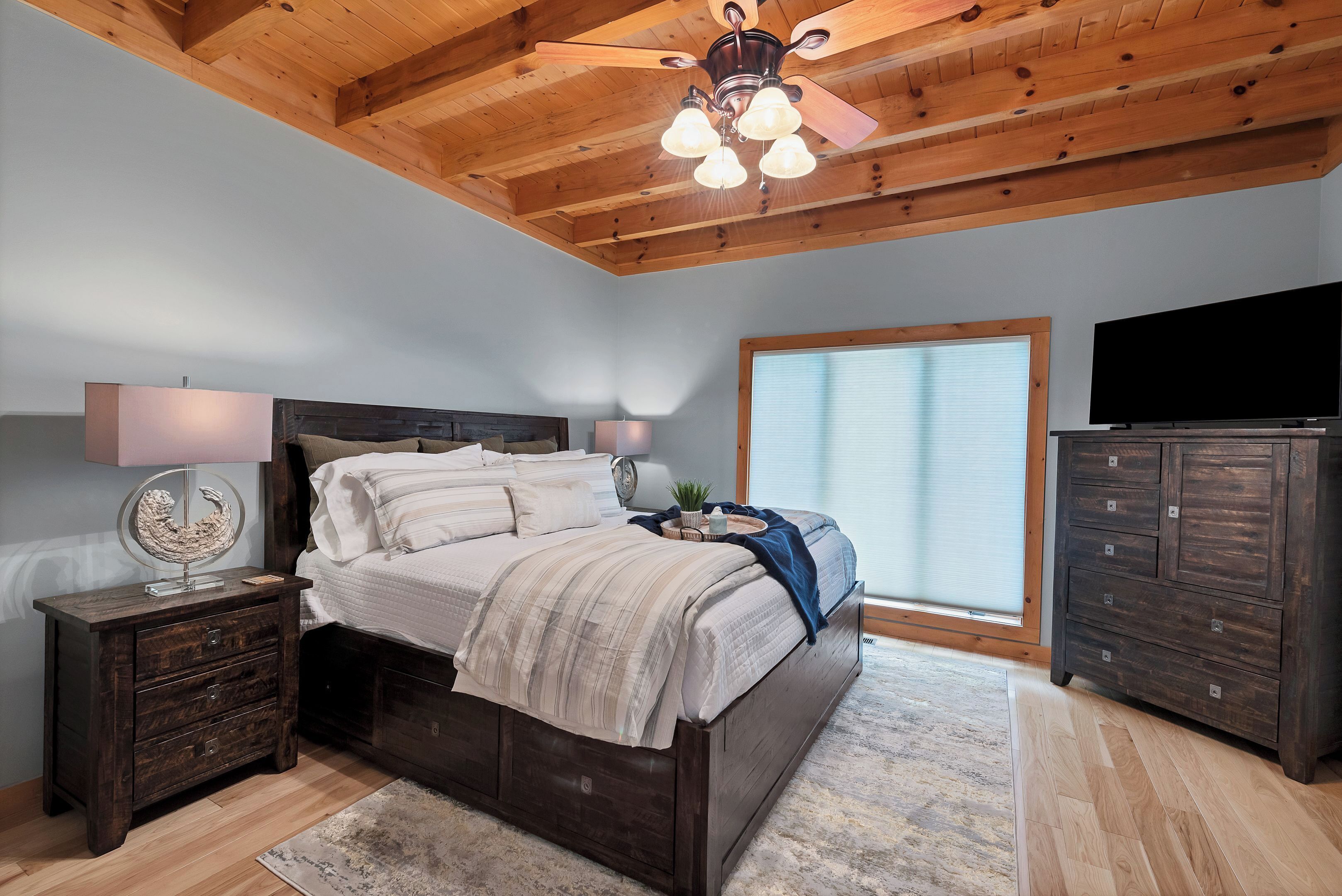 Une chambre avec un plafond en bois, un lit recouvert d’une couverture à rayures blanches et grises, une table de chevet avec des tiroirs, une commode et un ventilateur de plafond.