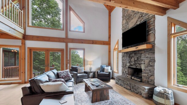 A spacious living room with a stone fireplace, wooden beams, large windows, and a cozy seating area. - This Amazing home has much to offer ,spacious rooms & and a hot-tub for relaxing (Townsend)