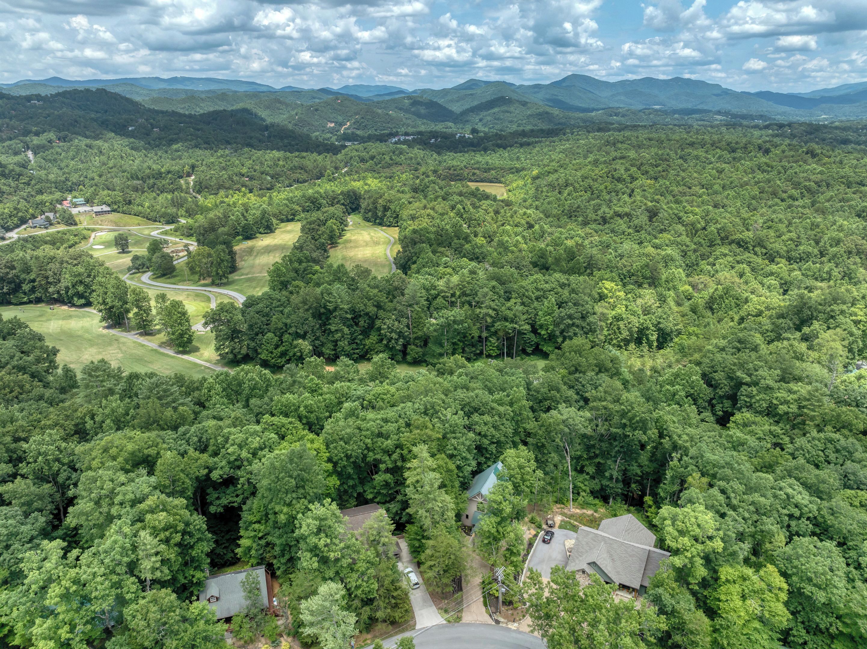 An aerial view of a residential area with houses surrounded by dense forest, a golf course, and rolling hills in the background.