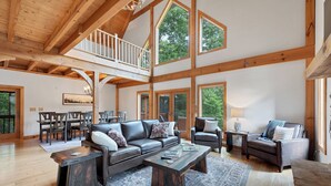 A spacious living room with a high ceiling, wooden beams, a balcony, and large windows. - This Amazing home has much to offer ,spacious rooms & and a hot-tub for relaxing (Townsend)