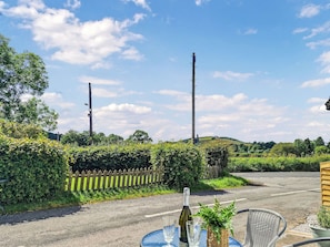 A roadside table with a bottle and glasses, surrounded by greenery and a wooden fence.