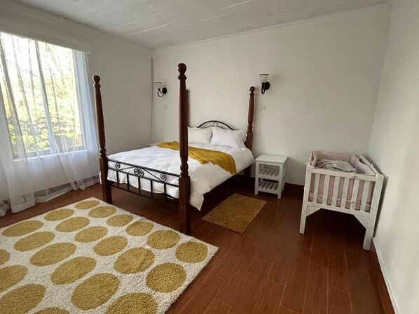 A bedroom with a four-poster bed, a crib, a bedside table, and a large window with curtains. - Enkishon Suites - 1 
 (Kiambu County)