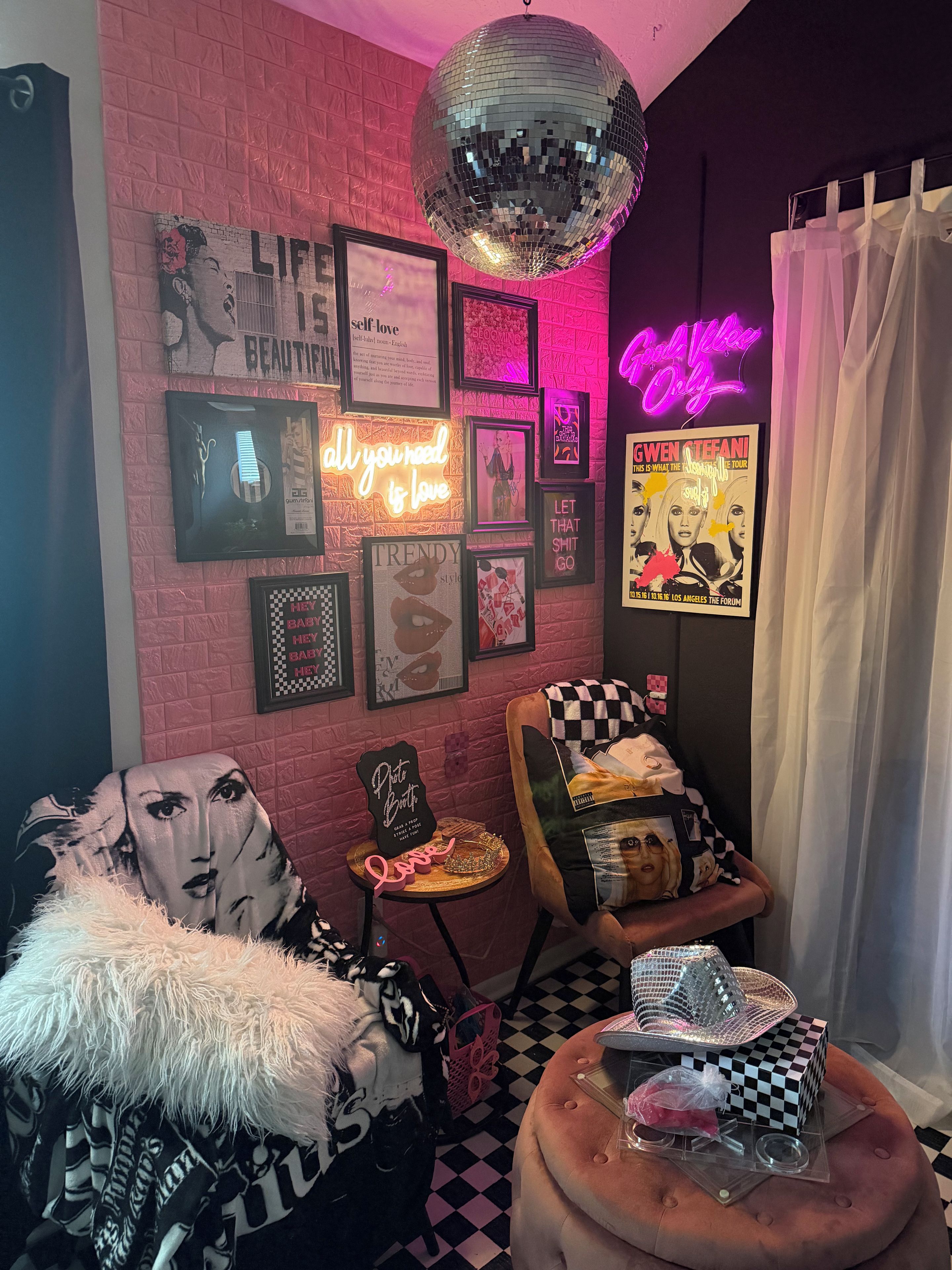 Una habitación con pared de ladrillo rosa, carteles enmarcados, un letrero de neón, una bola de discoteca y un reposapiés a cuadros.
