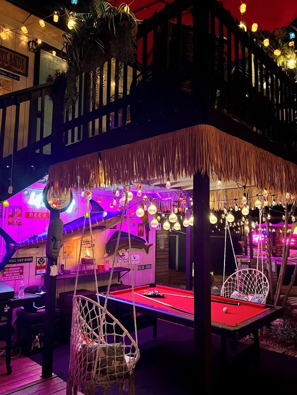A pool table with hanging chairs, a neon sign, and a tiki bar area.