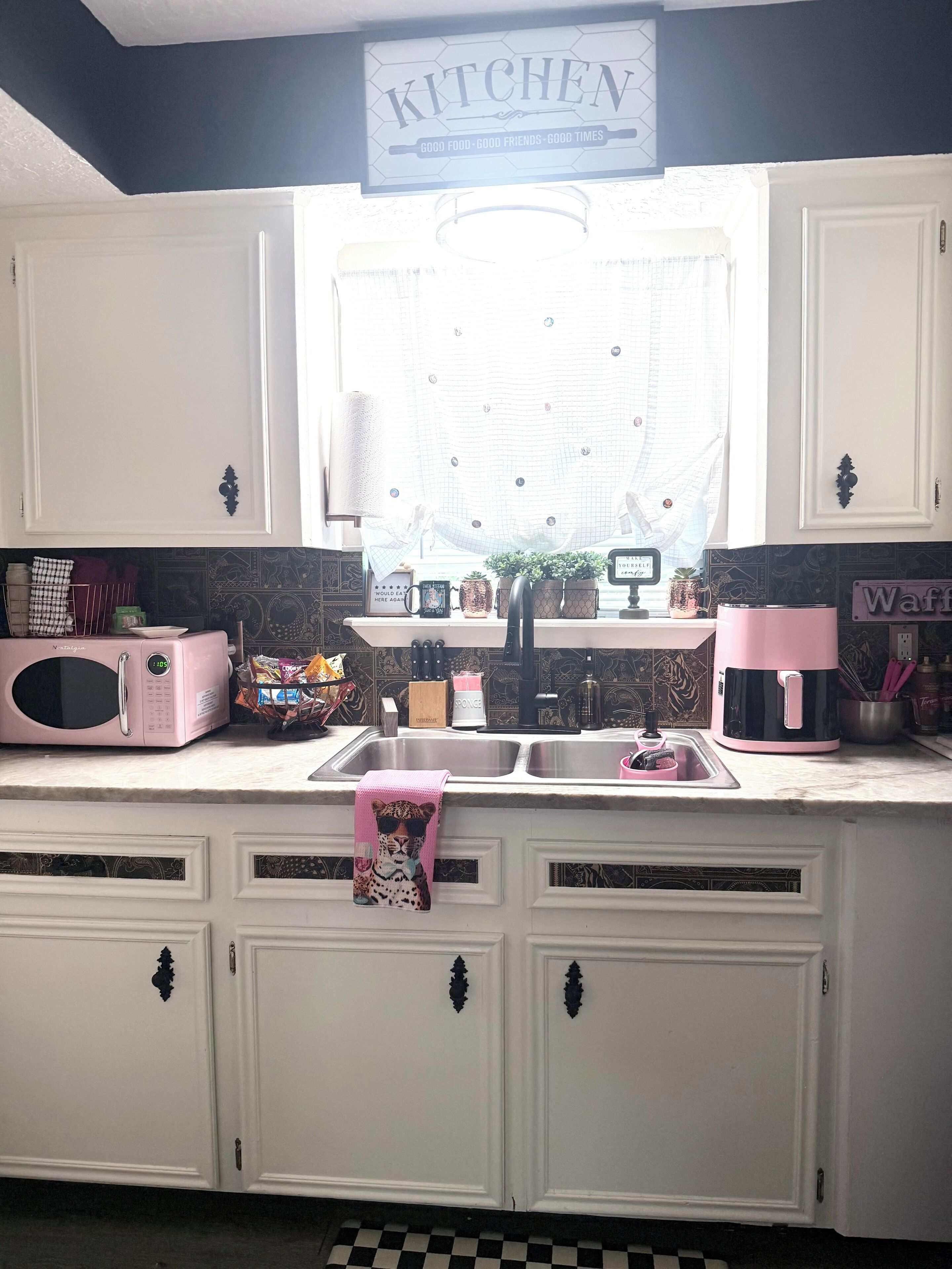 Una cocina con armarios blancos, microondas, pileta y ventana con cortina.