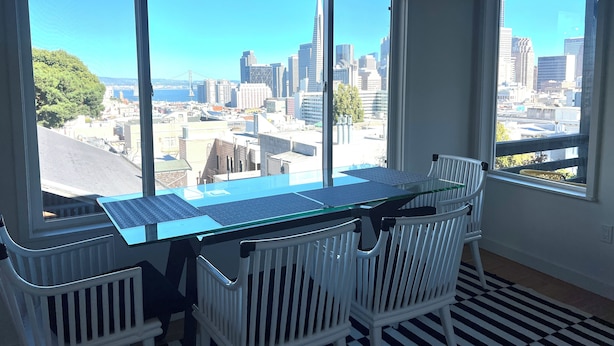 Une pièce avec une table à plateau de verre et des chaises blanches, de grandes fenêtres donnant sur la ville et un sol à rayures noires et blanches.