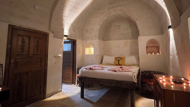 A bedroom with a bed, a wooden door, a bench, a table with candles, and a patterned rug. - Uluyol Stone House (Avanos)