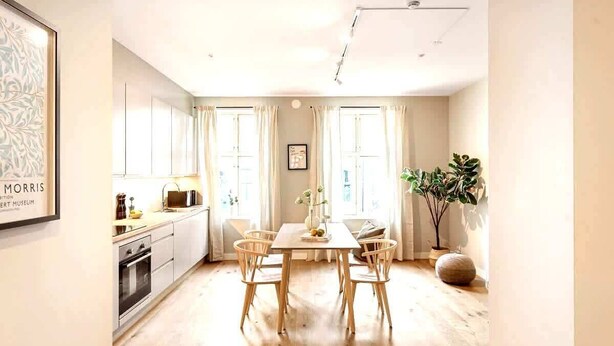 Une cuisine moderne et lumineuse, avec des meubles en bois, une table à manger entourée de chaises et une grande fenêtre agrémentée de rideaux.