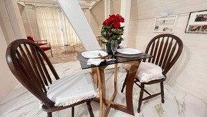A dining area with a glass table, two wooden chairs, a vase with red flowers, and a white curtain in the background. - Chalé Rústico 8 min do centro turístico (Campos do Jordão)