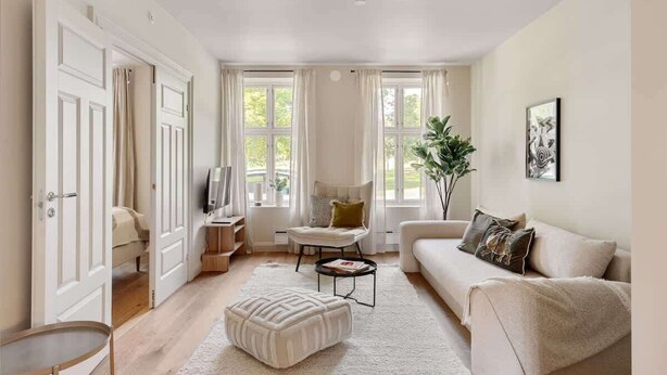 Un salon spacieux avec un canapé, un pouf et une table basse. On y trouve un téléviseur posé sur un meuble, une plante et une fenêtre avec des rideaux.