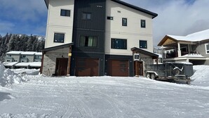 A multi-story residential building with a modern design, featuring a combination of white and dark siding, multiple windows, and a garage.