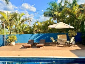 Área da piscina com espreguiçadeiras, uma mesa e um guarda-sol.