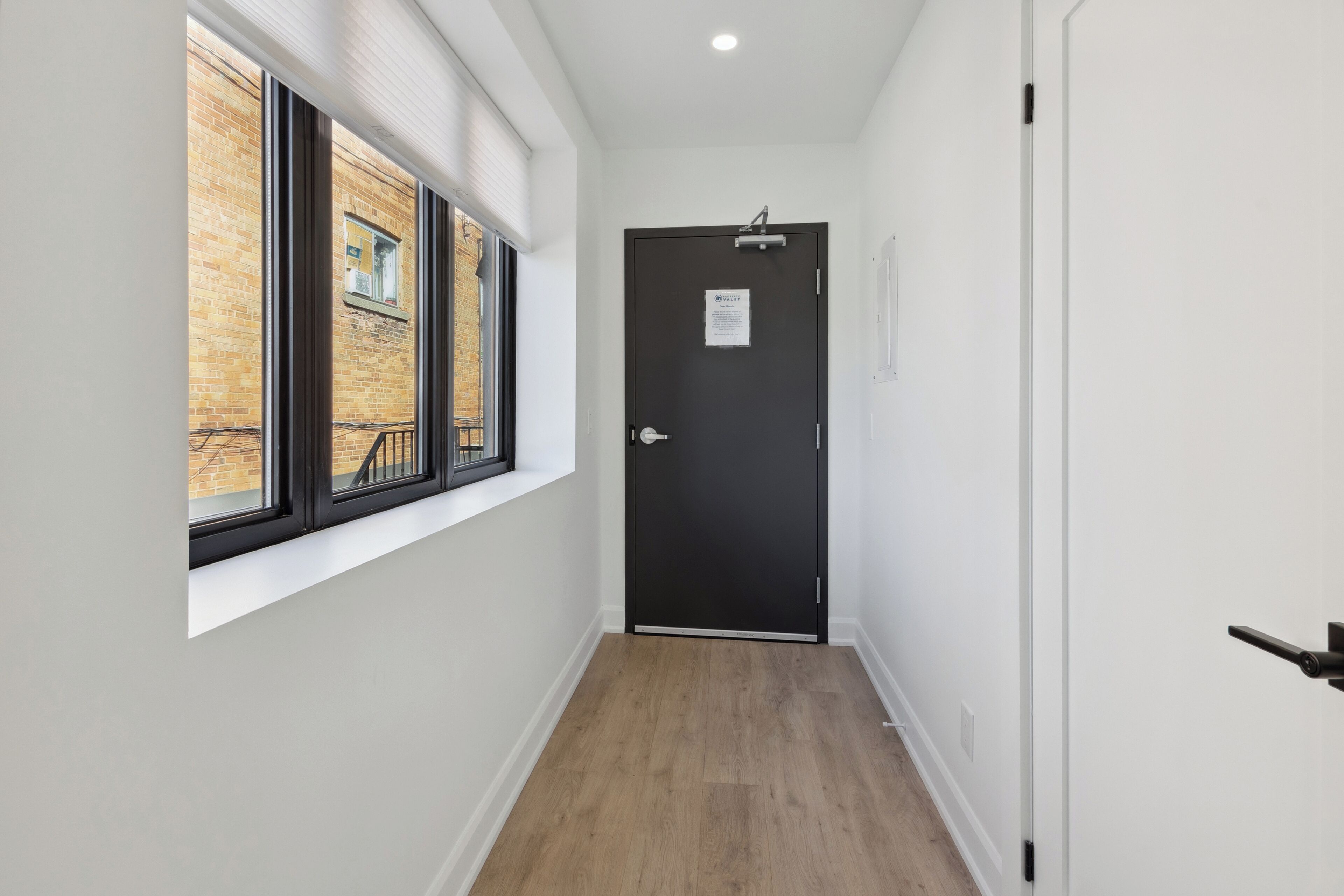 A narrow hallway with a black door, a window with blinds, and a wooden floor.
