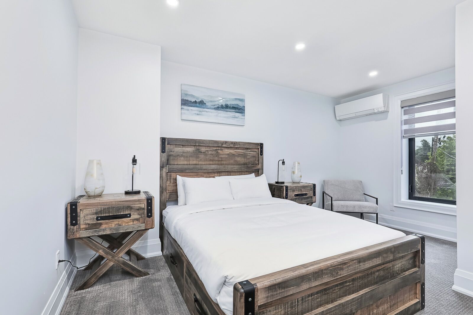 Un dormitorio con estructura de cama de madera, ropa de cama blanca, una mesita de noche con lámpara, una silla y una ventana con persianas.