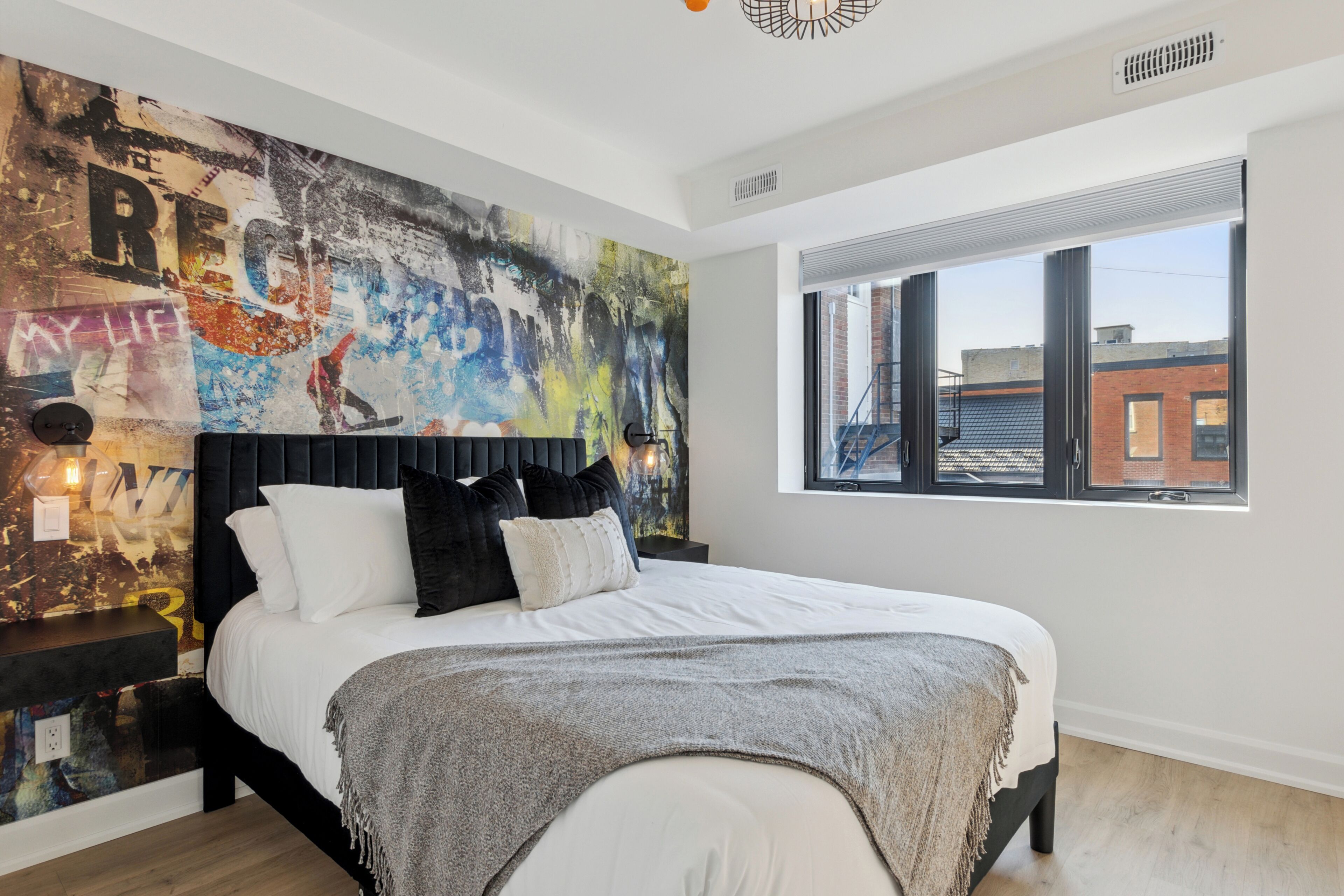 Une chambre moderne avec un grand lit, une fresque représentant un paysage urbain sur le mur et une fenêtre donnant sur des immeubles.