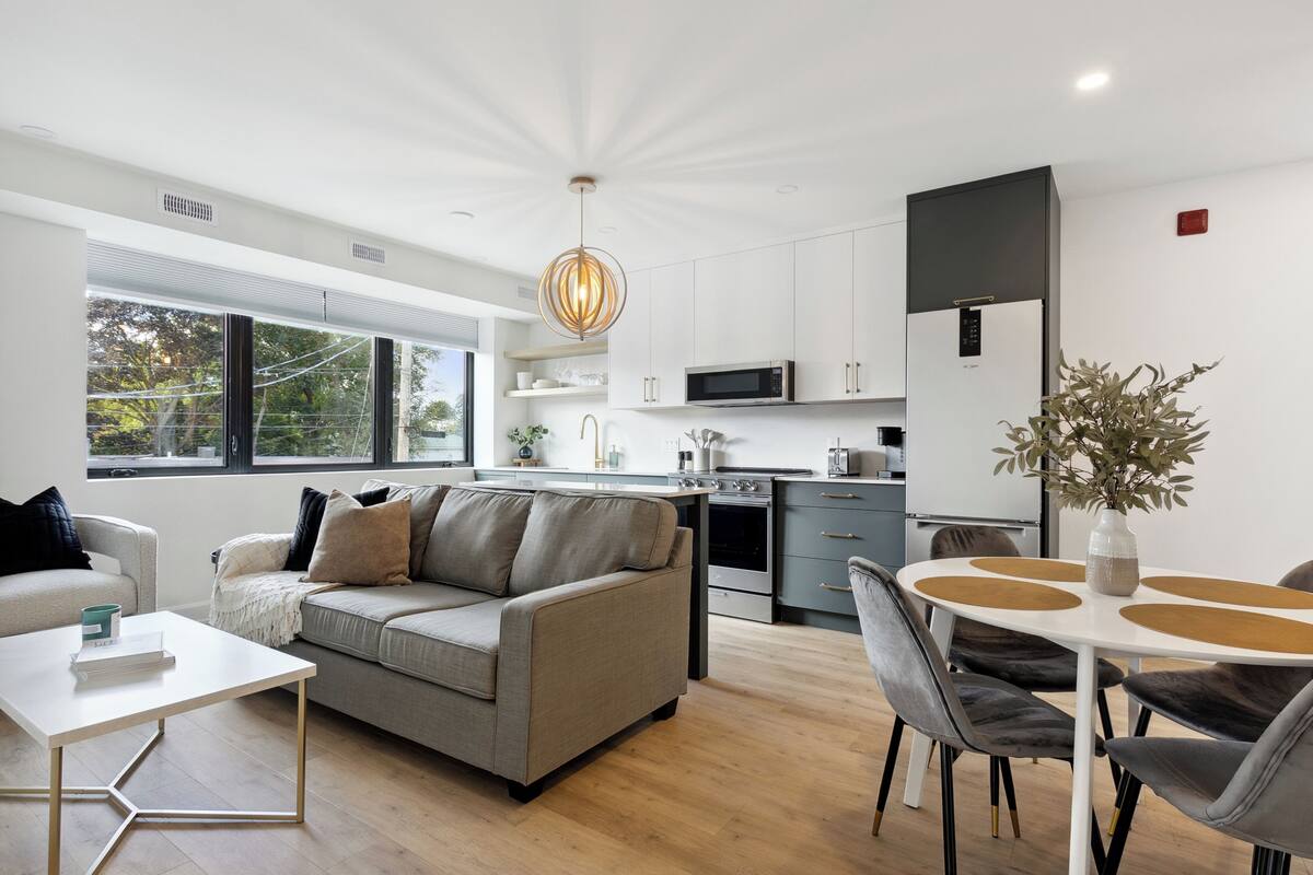Una sala de estar moderna con un sofá, una mesa de centro y un comedor con sillas. Se observa una cocina al fondo con un microondas y un horno.