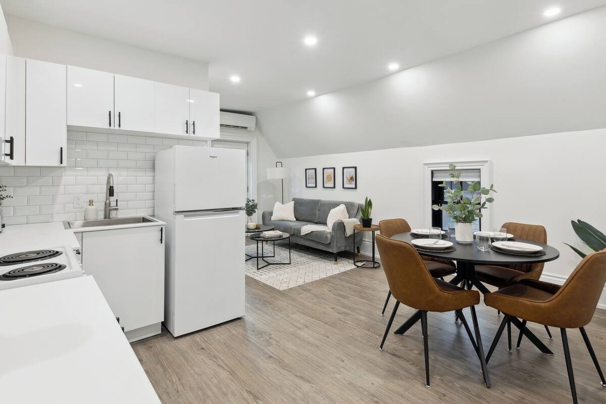 Una cocina moderna con armarios blancos, estufa integrada y refrigerador. Hay una zona de comedor con una mesa redonda y sillas marrones.