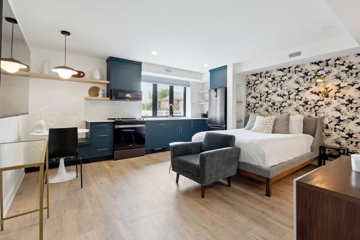 Un dormitorio moderno con una cama grande, un escritorio y una silla. Cuenta con una zona de cocina con armarios azules y un ventanal.