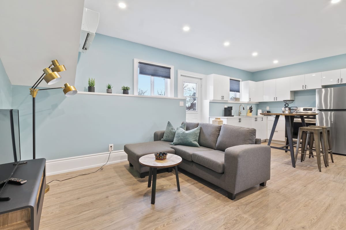 Un apartamento moderno con sofá gris, una pequeña mesa redonda, cocina con armarios blancos y un refrigerador de acero inoxidable.