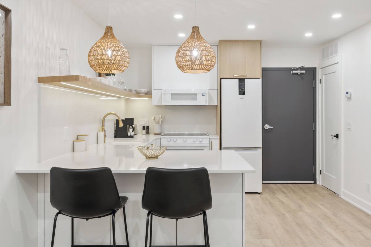 Una cocina moderna con armarios blancos, una isla central con dos sillas negras, un refrigerador y un microondas.