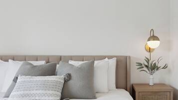 Una cama bien hecha con varios almohadones y una mesita de noche con una lámpara y un florero.