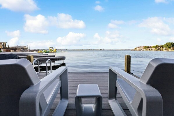 Una terraza de madera con barandillas metálicas y una pequeña mesa, con vista a aguas tranquilas y edificios lejanos.