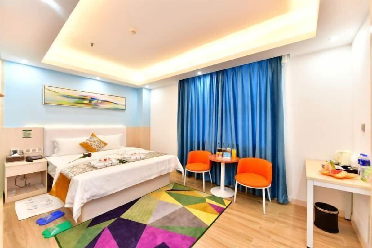 A hotel room with a bed, two orange chairs, a small table, a desk, and a colorful rug.