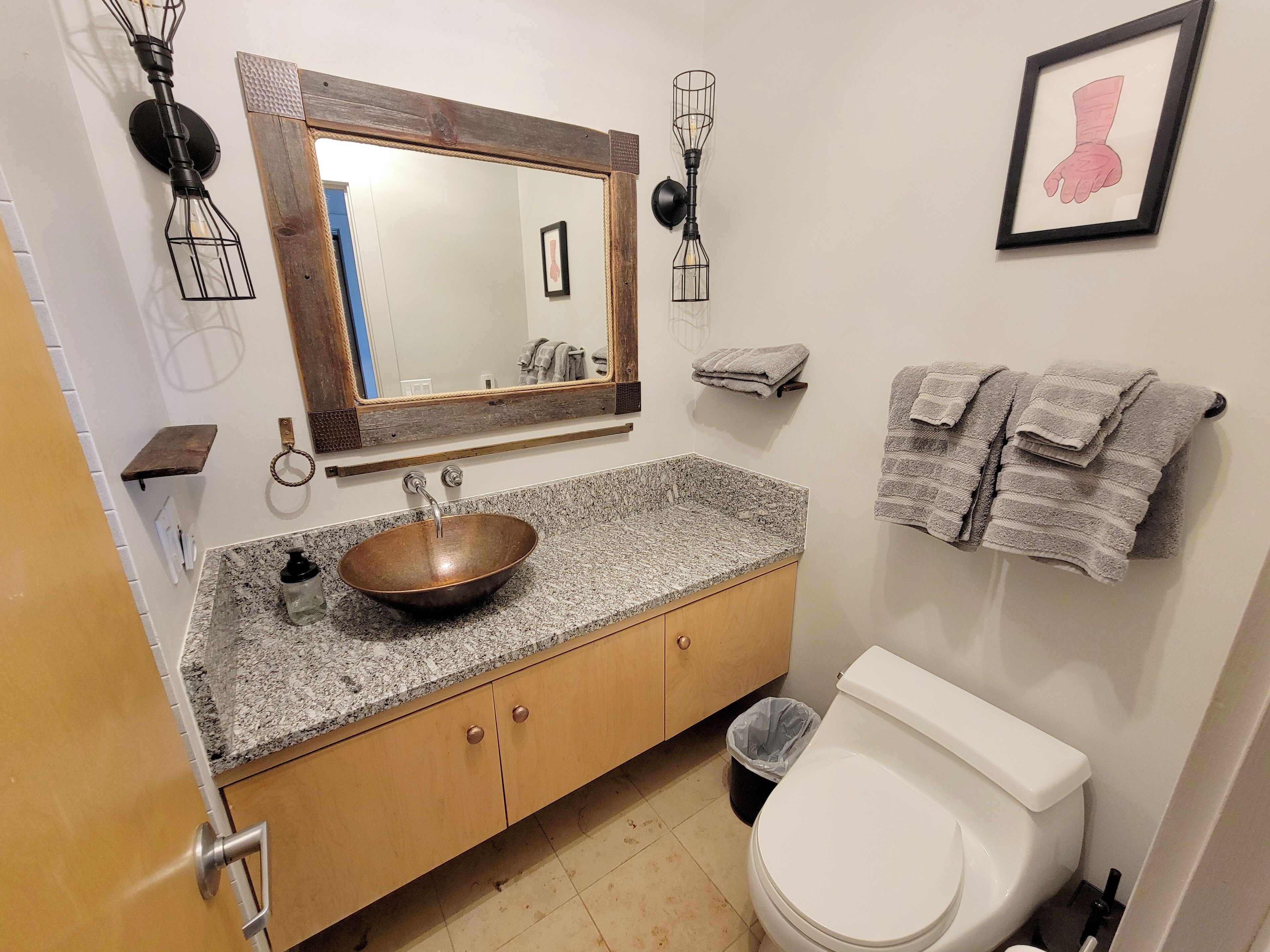 Une salle de bain avec un meuble-lavabo en bois, un évier en cuivre, des toilettes blanches et une œuvre d’art encadrée au mur.