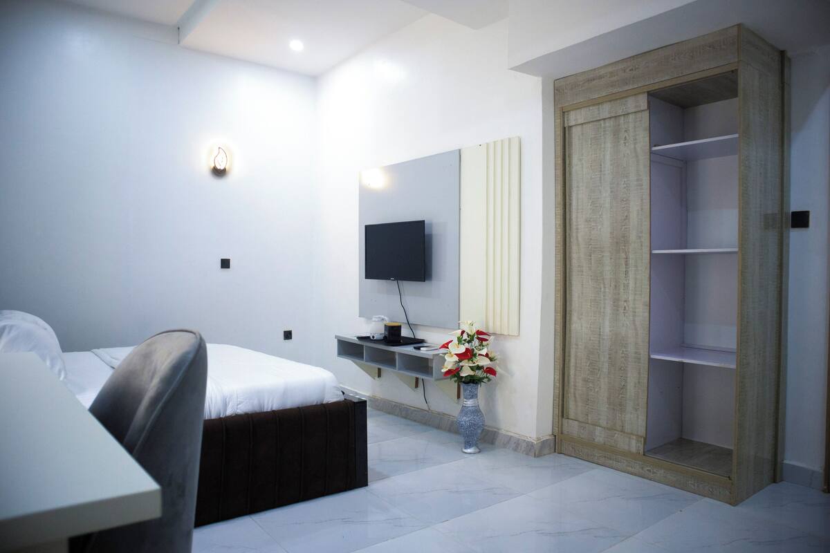 A modern hotel room with a bed, a desk, a television, a vase with flowers, and a wardrobe.