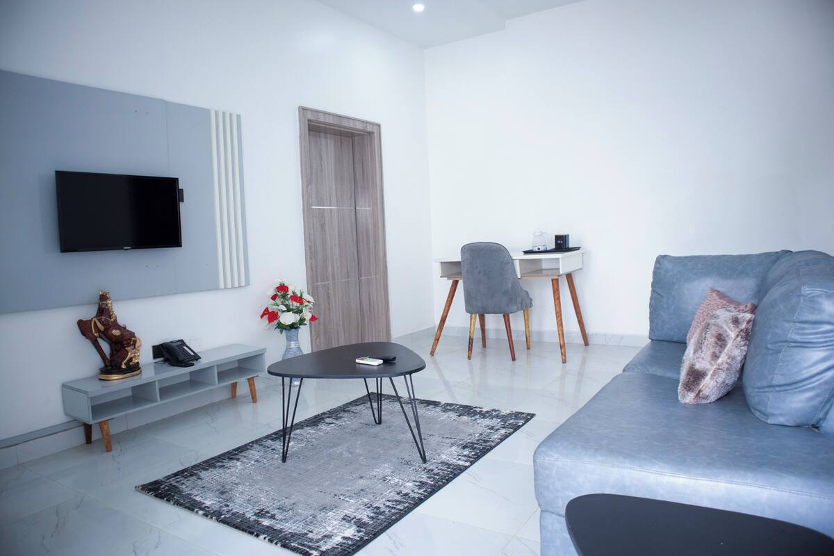 A modern living room with a grey sofa, a black coffee table, a TV mounted on the wall, a small desk with a chair, and a decorative statue.