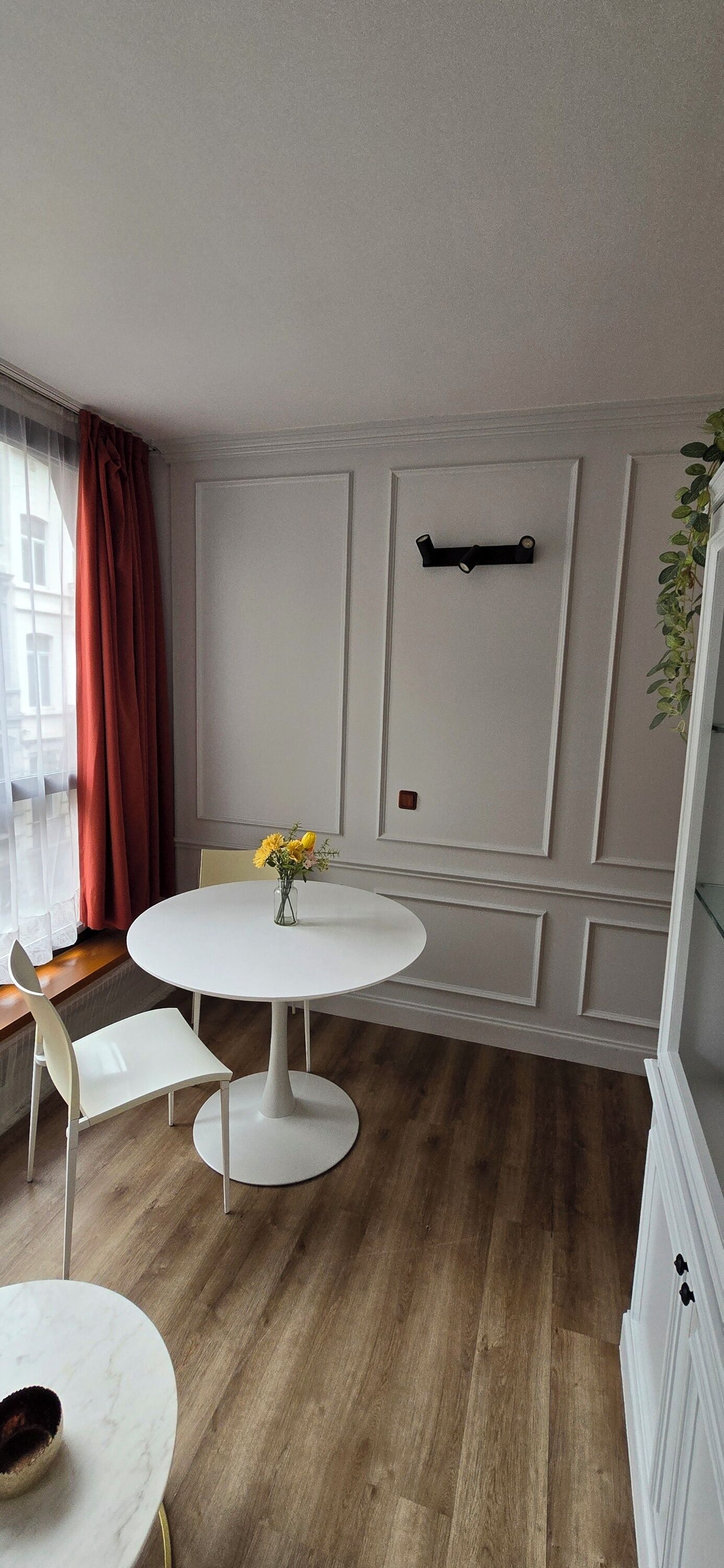 A dining area with a round white table, two white chairs, a vase with yellow flowers, and a wooden floor.