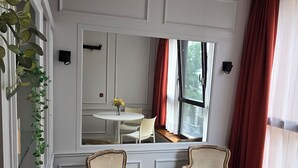 Una stanza con due poltrone, un tavolino rotondo e vista su una sala da pranzo attraverso una finestra con tende rosse.