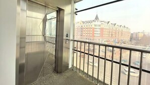 Un balcón con baranda metálica, vista de un paisaje urbano con edificios y una puerta con panel de vidrio.
