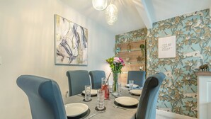 Un coin repas avec une table dressée pour quatre personnes, des chaises bleues, un vase de fleurs et un mur orné d’un motif de feuilles tropicales.