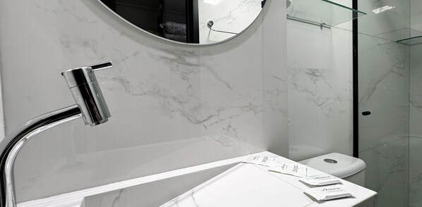 A modern bathroom with a marble countertop, a round mirror, and a glass shower enclosure. - MOVE (João Pessoa)