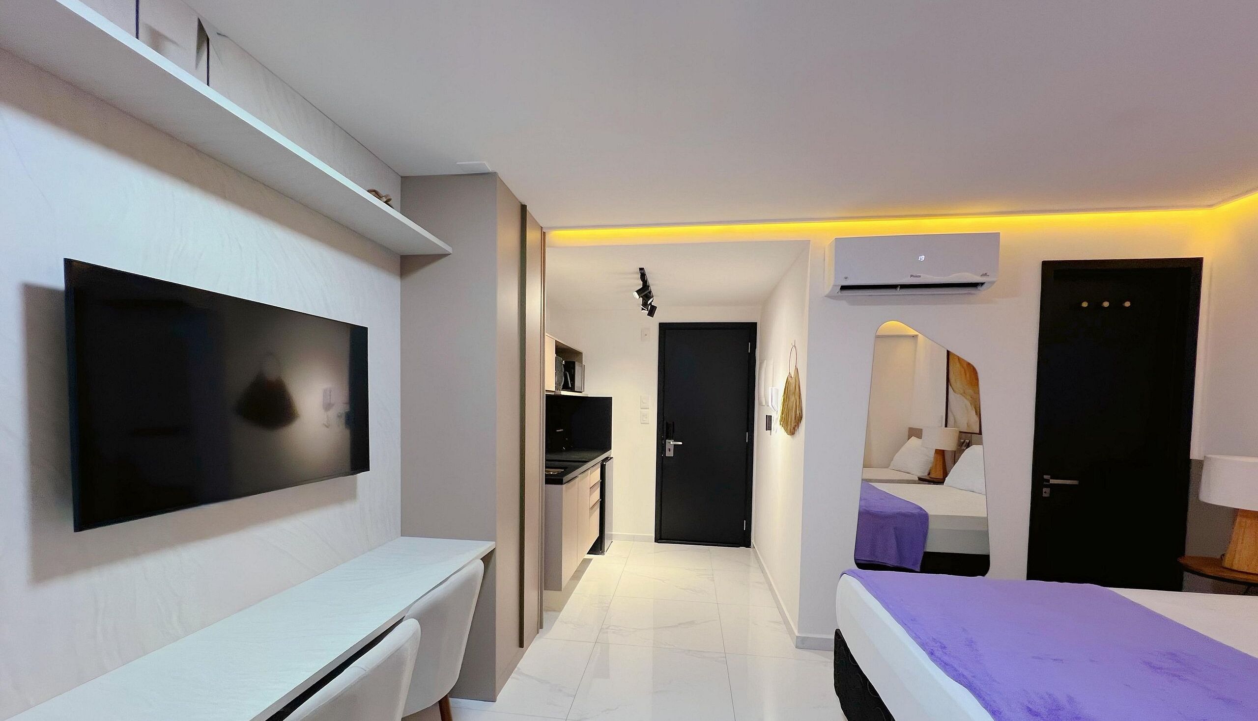 A modern hotel room with a flat-screen TV, a bed with a purple blanket, a desk, and a small kitchenette.