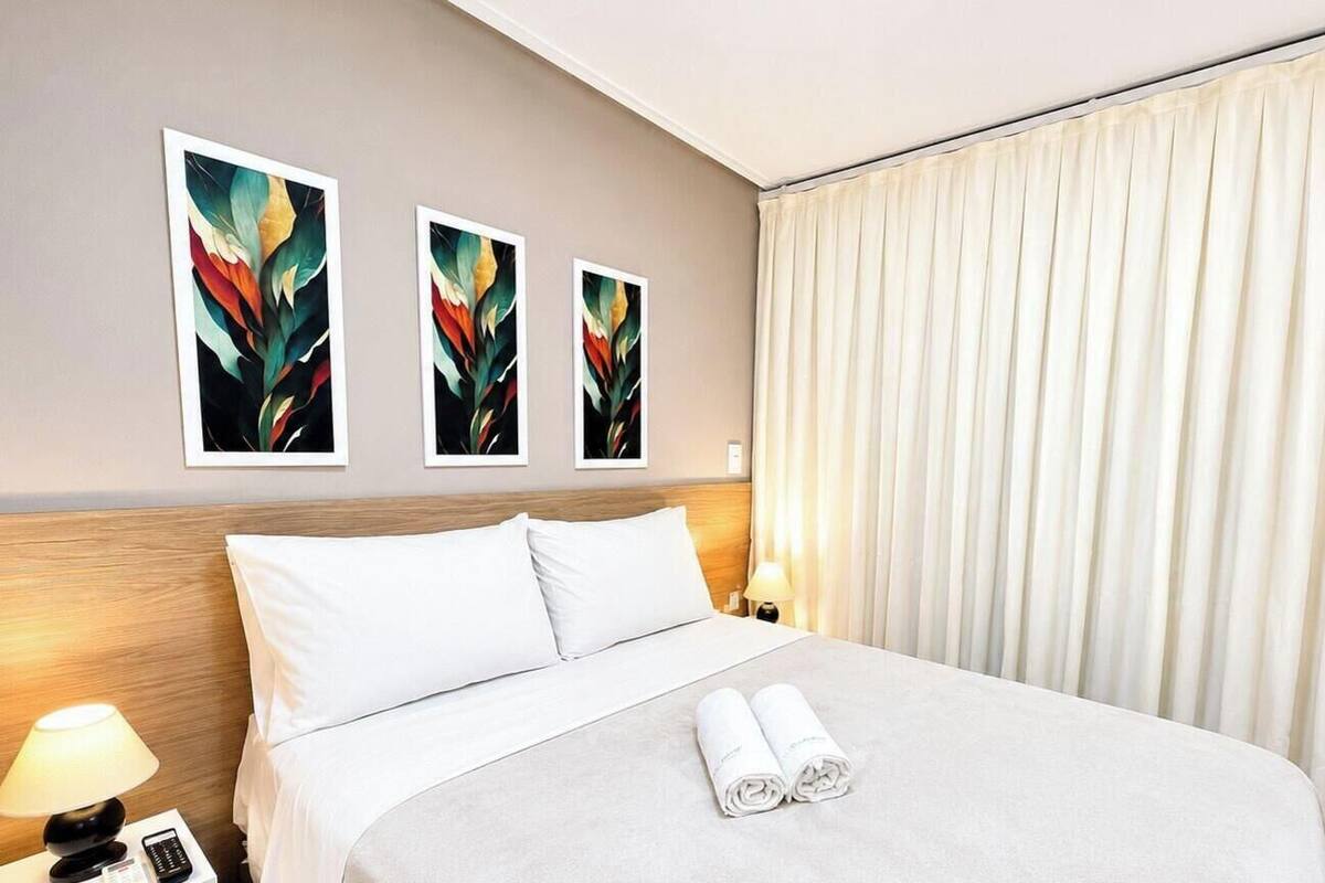 A hotel room with a bed, white pillows, a lamp, a remote control, and three framed artworks on the wall.