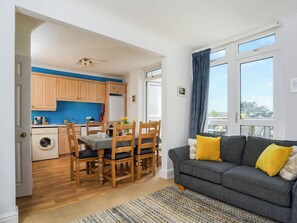 A living room with a grey sofa and yellow cushions, a dining area with wooden chairs and a table, and a kitchen with wooden cabinets and a blue accent wall.