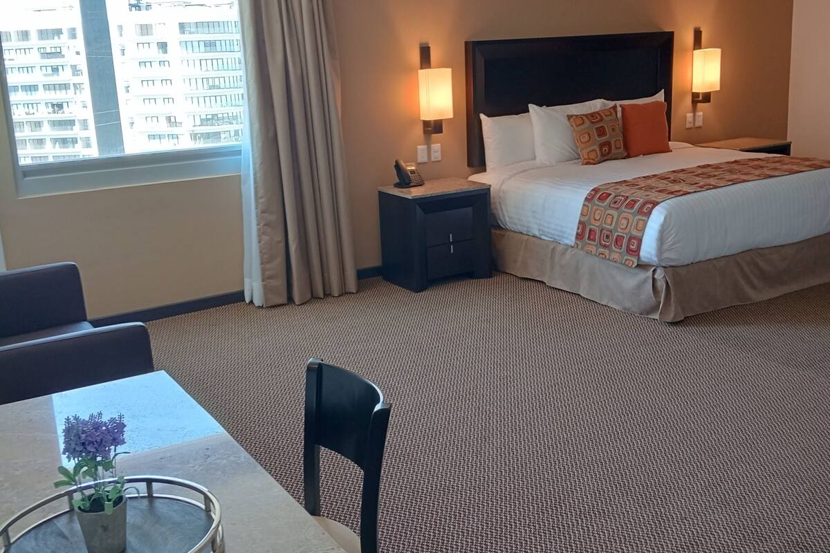 A hotel room with a bed, bedside tables, a desk with a chair, and a view of buildings outside the window.