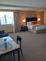 Habitación de hotel con cama, mesitas de noche, escritorio con silla y vistas a edificios desde la ventana.