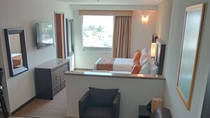 A compact living space with a sofa, a small table, a potted plant, a window with curtains, a bed, and a TV mounted on the wall. - Ankara Pedregal Centro de Convenciones (San Luis Potosi)