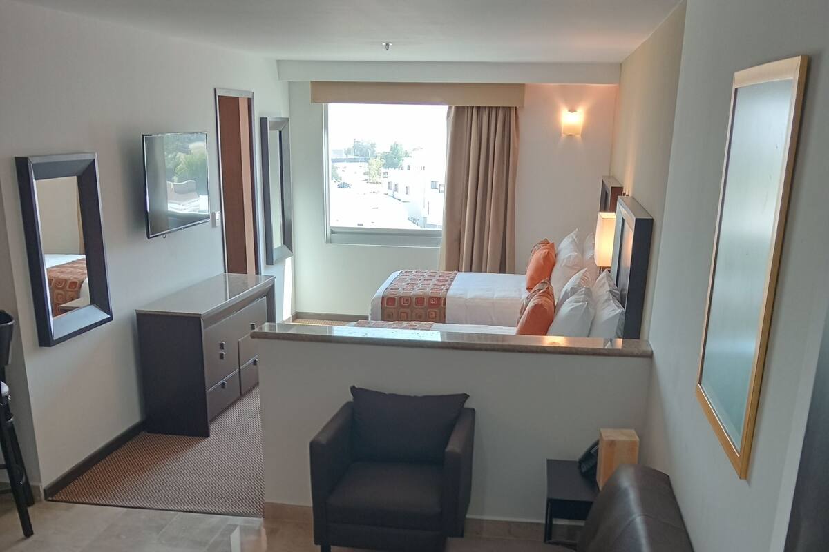 A compact living space with a sofa, a small table, a potted plant, a window with curtains, a bed, and a TV mounted on the wall.