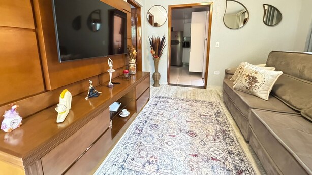 Een woonkamer met een grote flatscreen televisie, een houten tv-meubel, een kamerkleed met dessin en een bruine bank.